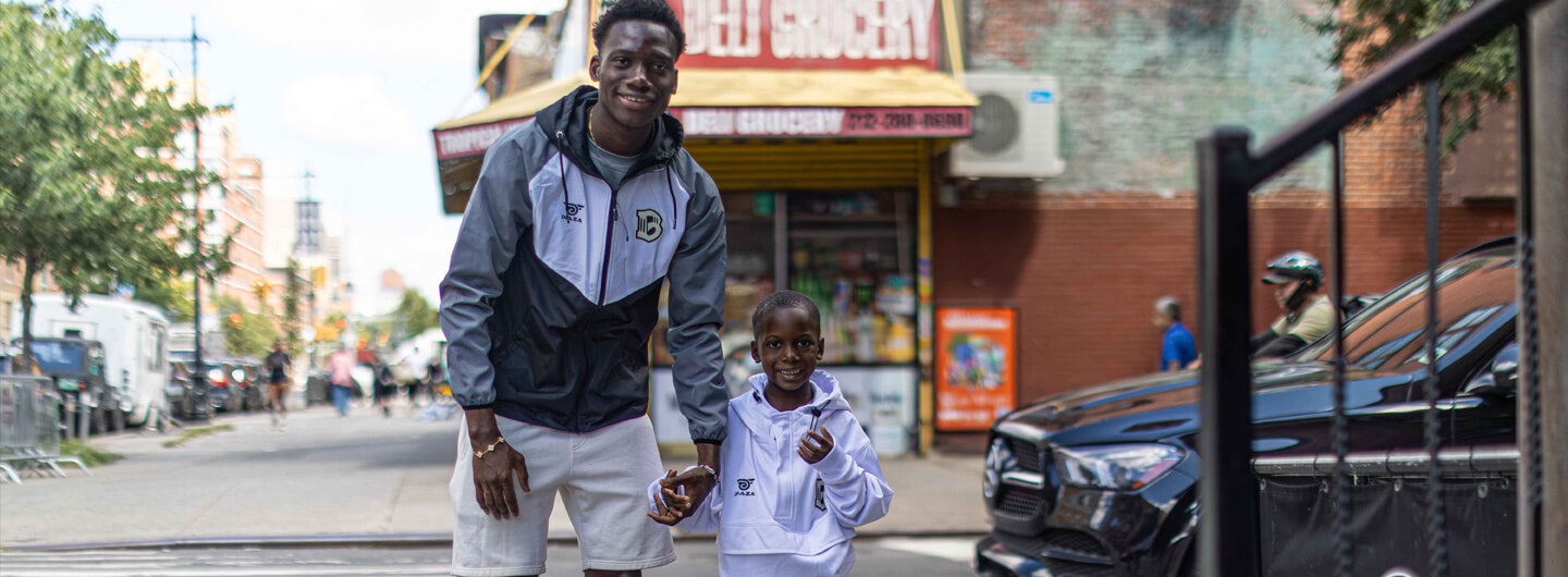 A man walks hand in hand with a boy across the street, while both wear Brooklyn FC clothing